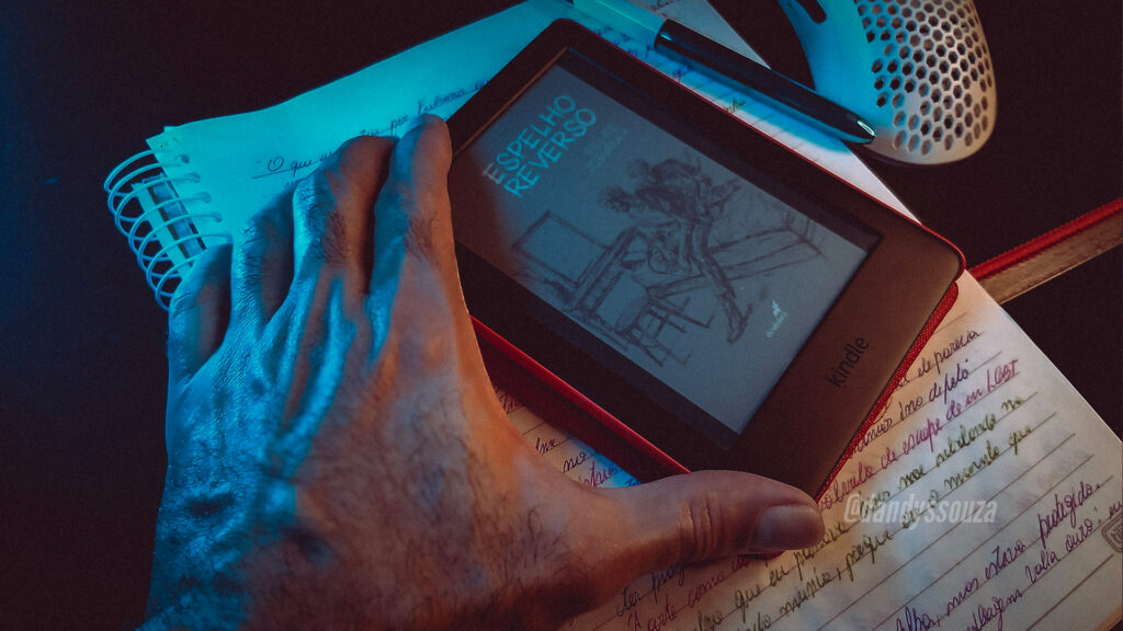 Espelho Reverso | Foto autoral do blog: uma mão segurando o livro no Kindle em cima do caderno com anotações. Cenário escuro com luz neon azul | Dandy Souza