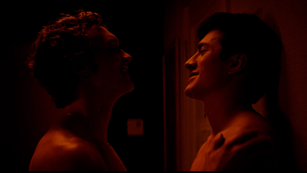 cena de Connor Storrie encostando Hudsson Willaams na parede para beijá-lo. Ocenário está vermelho e escuro | Dandy Souza